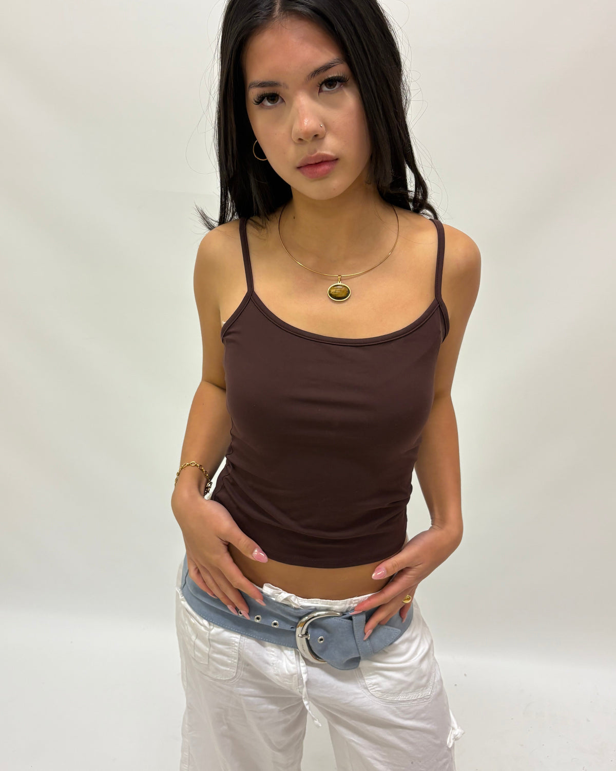 A woman modeling a dark brown spaghetti-strap camisole tank top paired with white pants and a blue belt, standing against a plain off-white studio background.