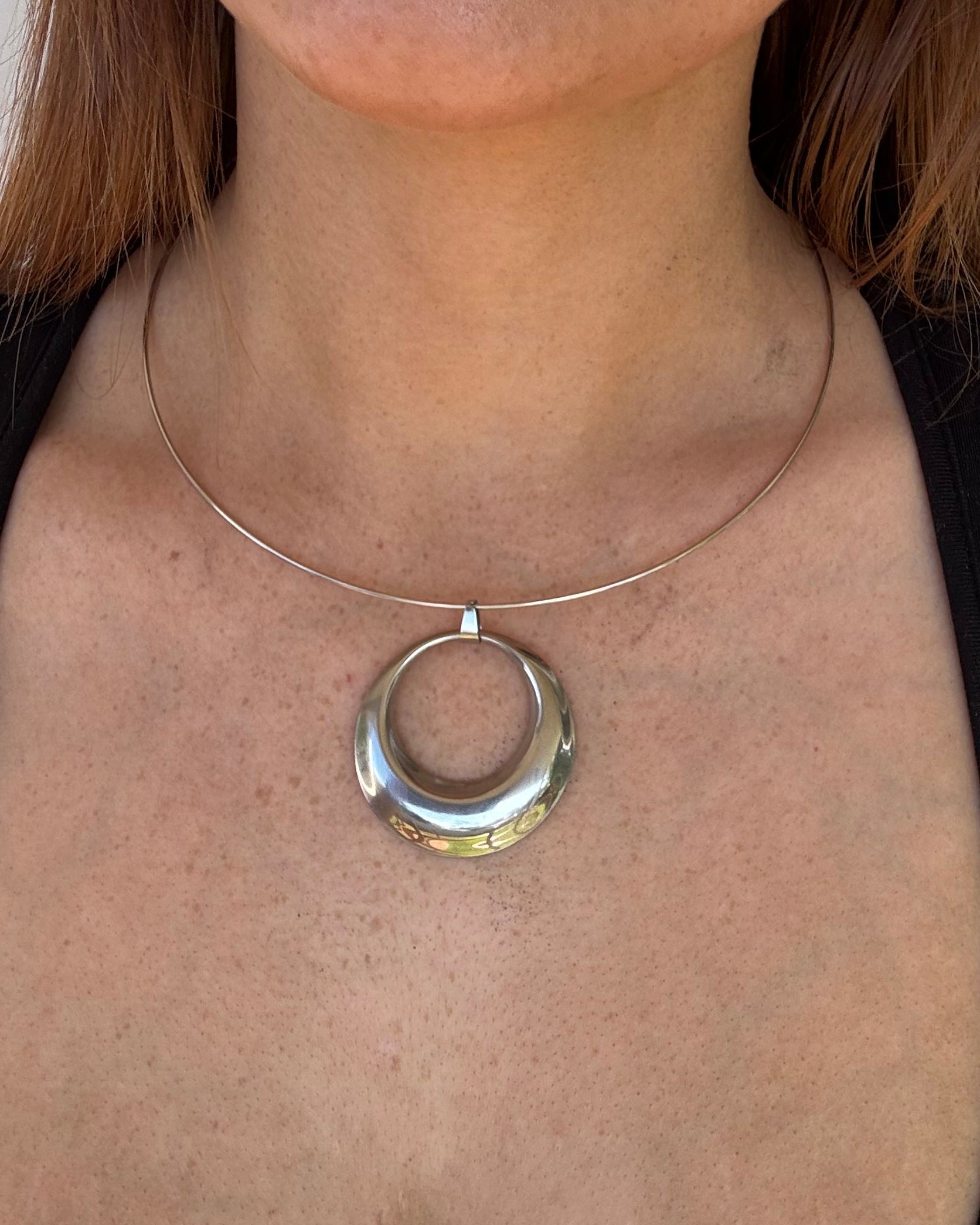 A close-up of a person lying down wearing a silver circular collar necklace with a thin wire loop extending from the center of the chest.